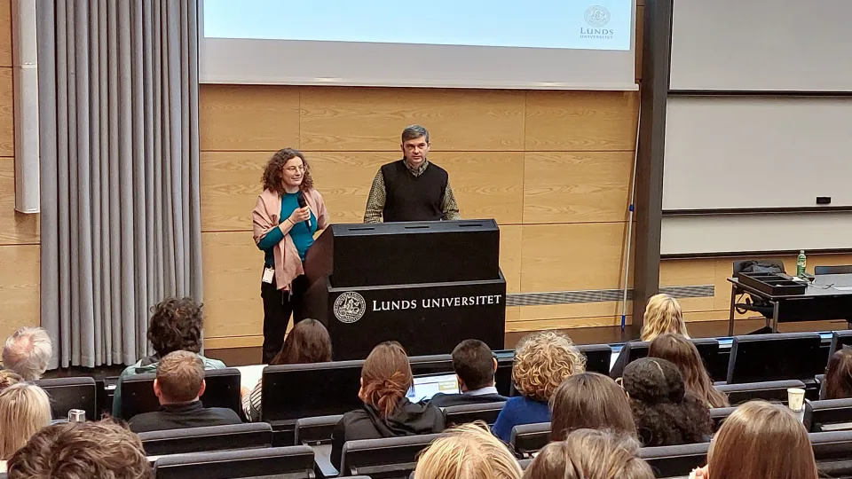 Two persons standing behind a podium in front of an audience.