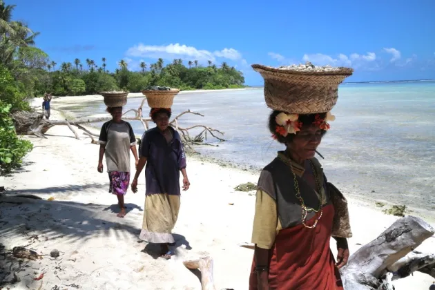 Three individuals walking on a beach while carrying baskets on their heads.